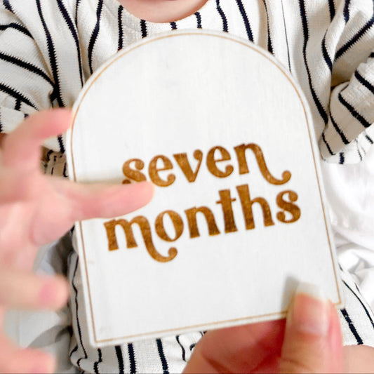 Baby in a striped outfit holding a 'seven months' milestone card against a white background
